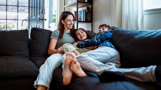 A woman is shown lying down on the couch with her two children. They are all laughing.