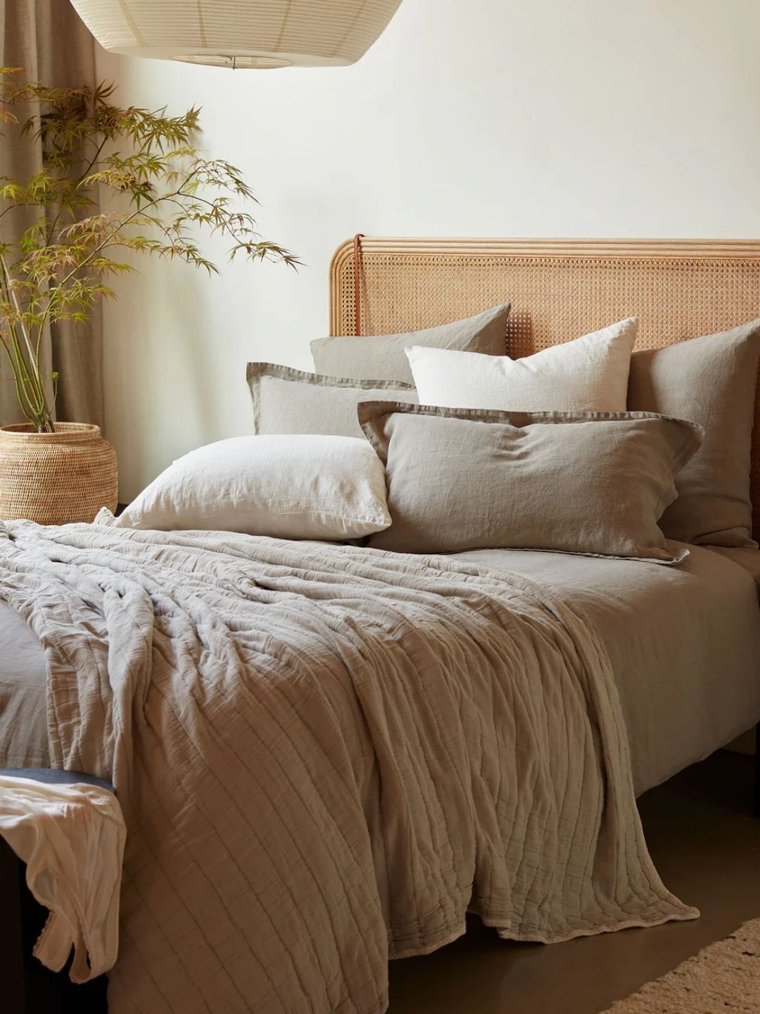 Image of a bed with a rattan headboard in relaxed, light gray linen bedding in a Wabi-Sabi style room. There are six pillows on the bed, a throw, and a potted plant in the corner.