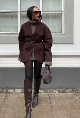 winter tight outfit shown in a photo of a woman wearing a brown blazer over a brown turtleneck sweater dress styled with a black belt, black sheer tights, brown knee-high boots, and a brown handbag