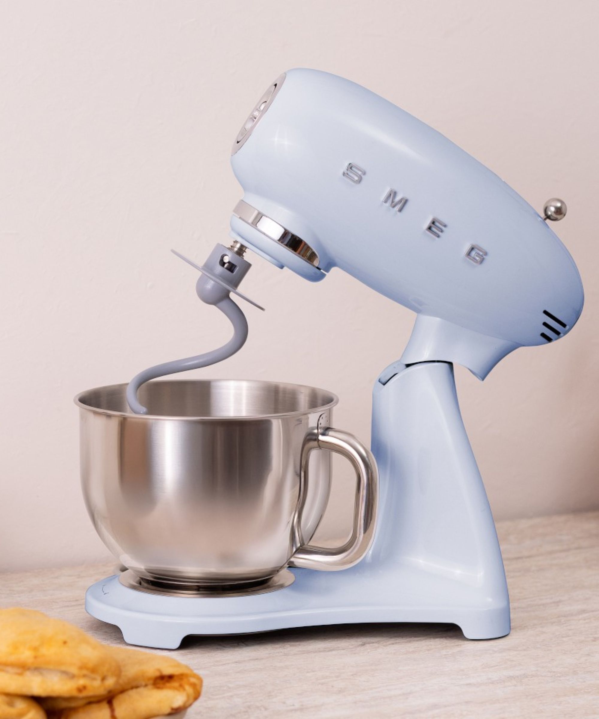A pastel blue stand mixer with the head tilted back, with a dough hook attachment in a stainless steel bowl. On a wooden counter against a beige wall.