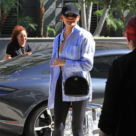 Chrissy Teigen wears black heeled ankle boots, black leggings, a striped button-down shirt, a black Gucci crossbody bag, a black hat, and white sports bra.