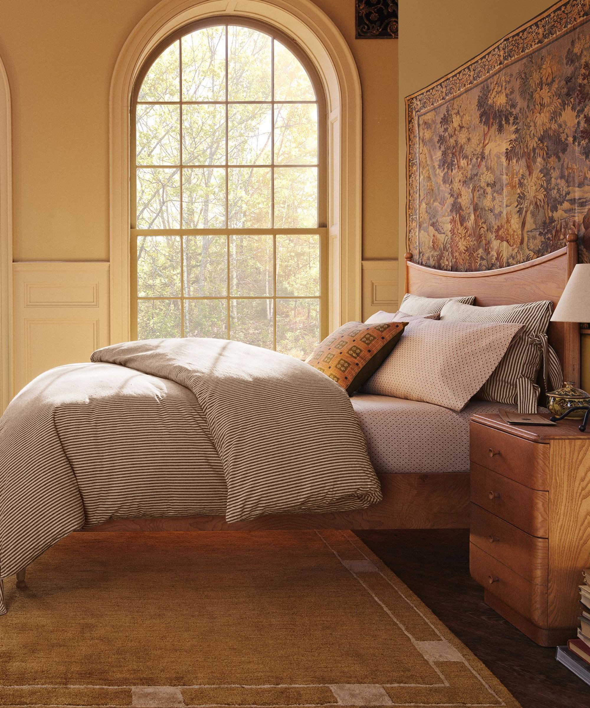 warm butter yellow bedroom with arched windows styled for the Pierce & Ward x West Elm collection with a wooden bed frame, tapestry hung on the wall, and a large ochre rug underneath