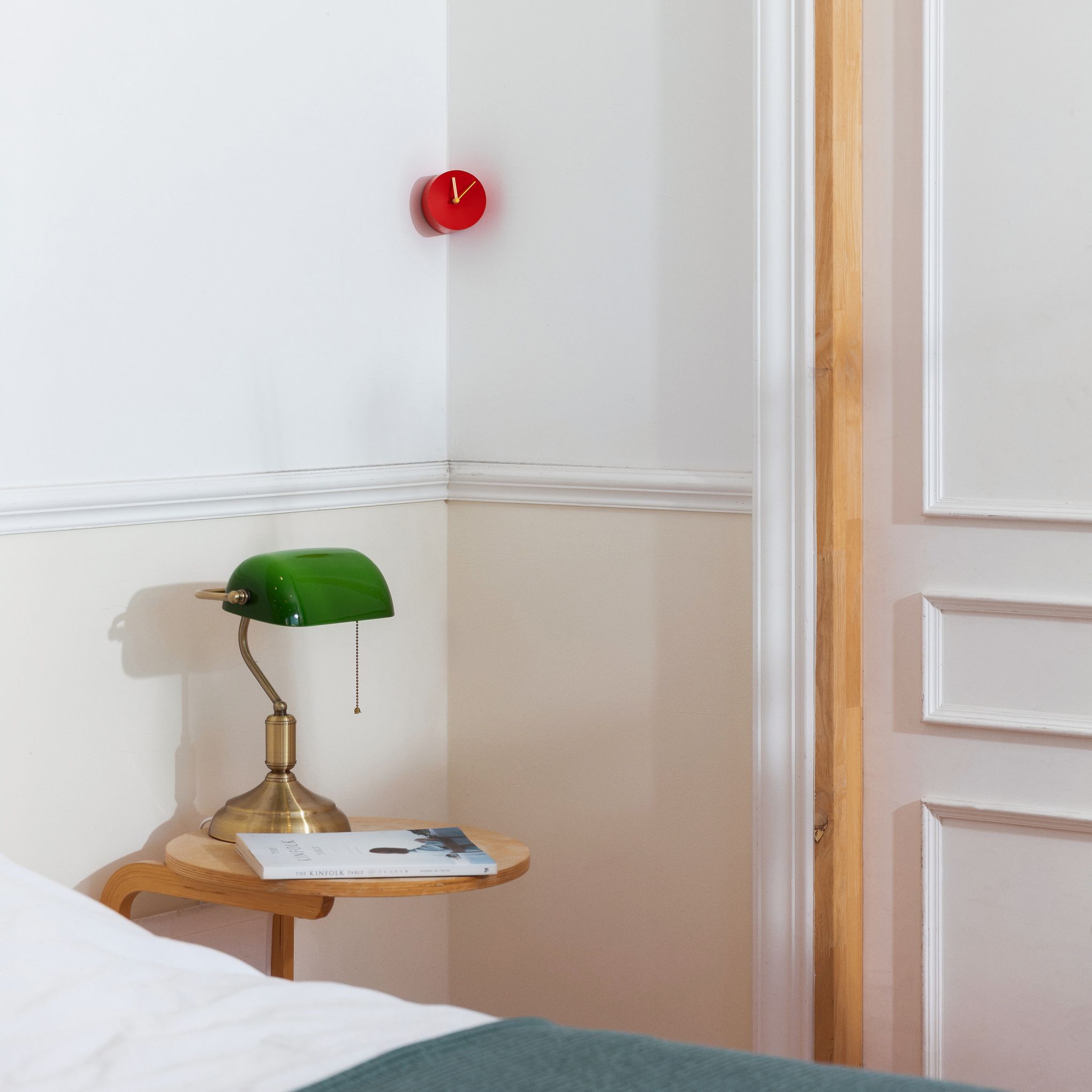 A round red clock hangs in the corner of a white wall above a bedside table with a green and brass desk lamp.