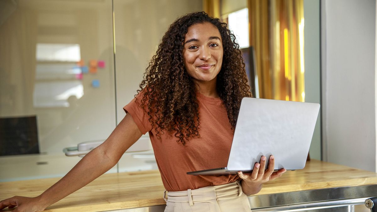 A young black woman holding a silver laptop and smiling at the camera