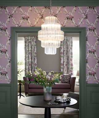 Hallway with gray-blue painted paneling, purple floral wallpaper, glass chandelier hanging above a black round wooden table with flowers, living room with floral curtains, purple sofa