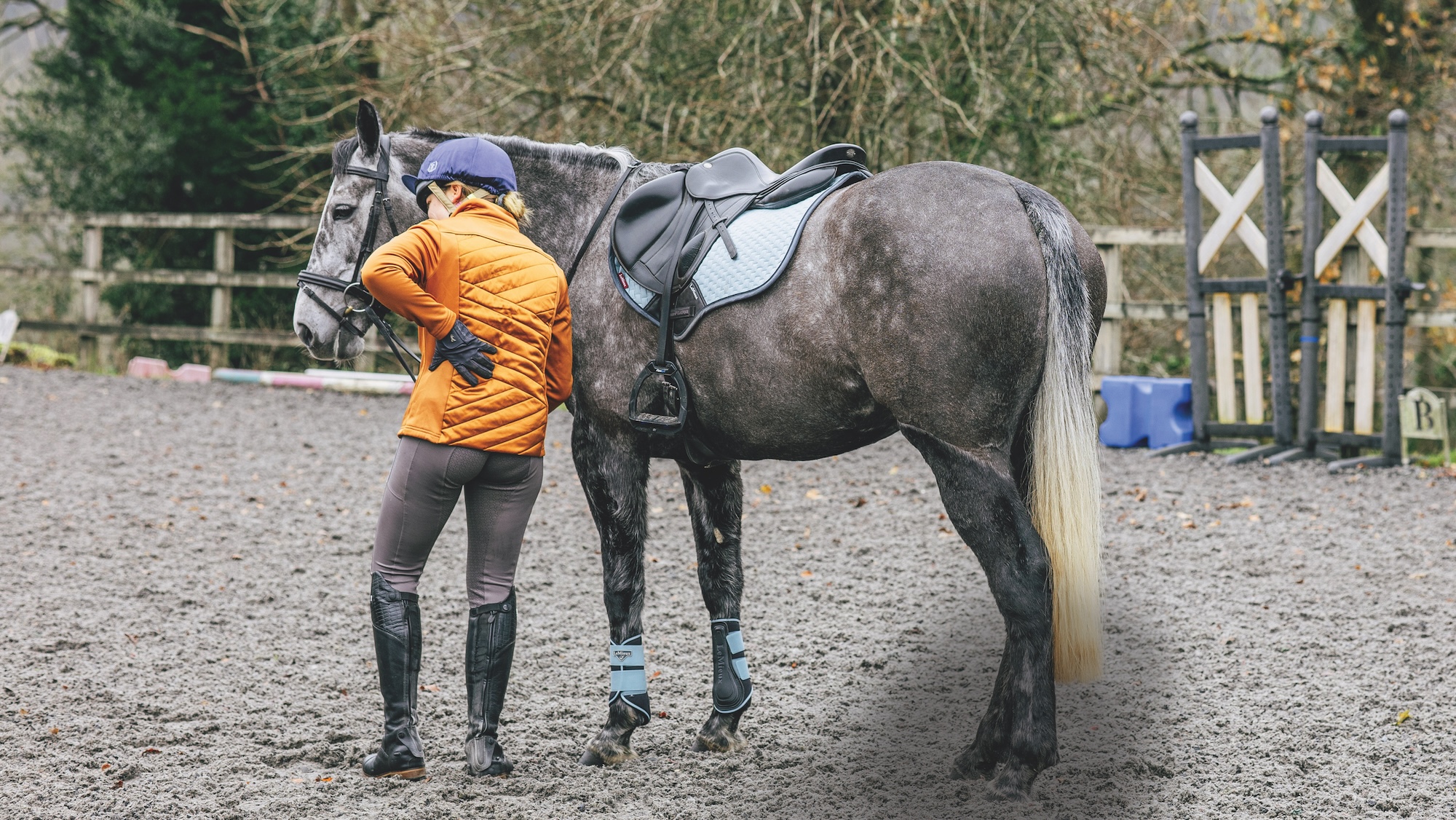 A rider in an orange jacket holds her lower back with her left arm. Next to her, a dark grey horse stands quietly. The pair are in an all-weather arena.
