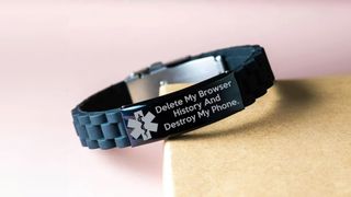 Medical alert bracelet with phrase "Delete My Browser History, and Destroy My Phone" etched into it placed on corner of a wooden surface against a light pink wall.