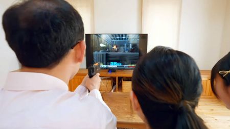 Family sitting in living room using G'aim'e Time Crisis Plug and Play console on TV.