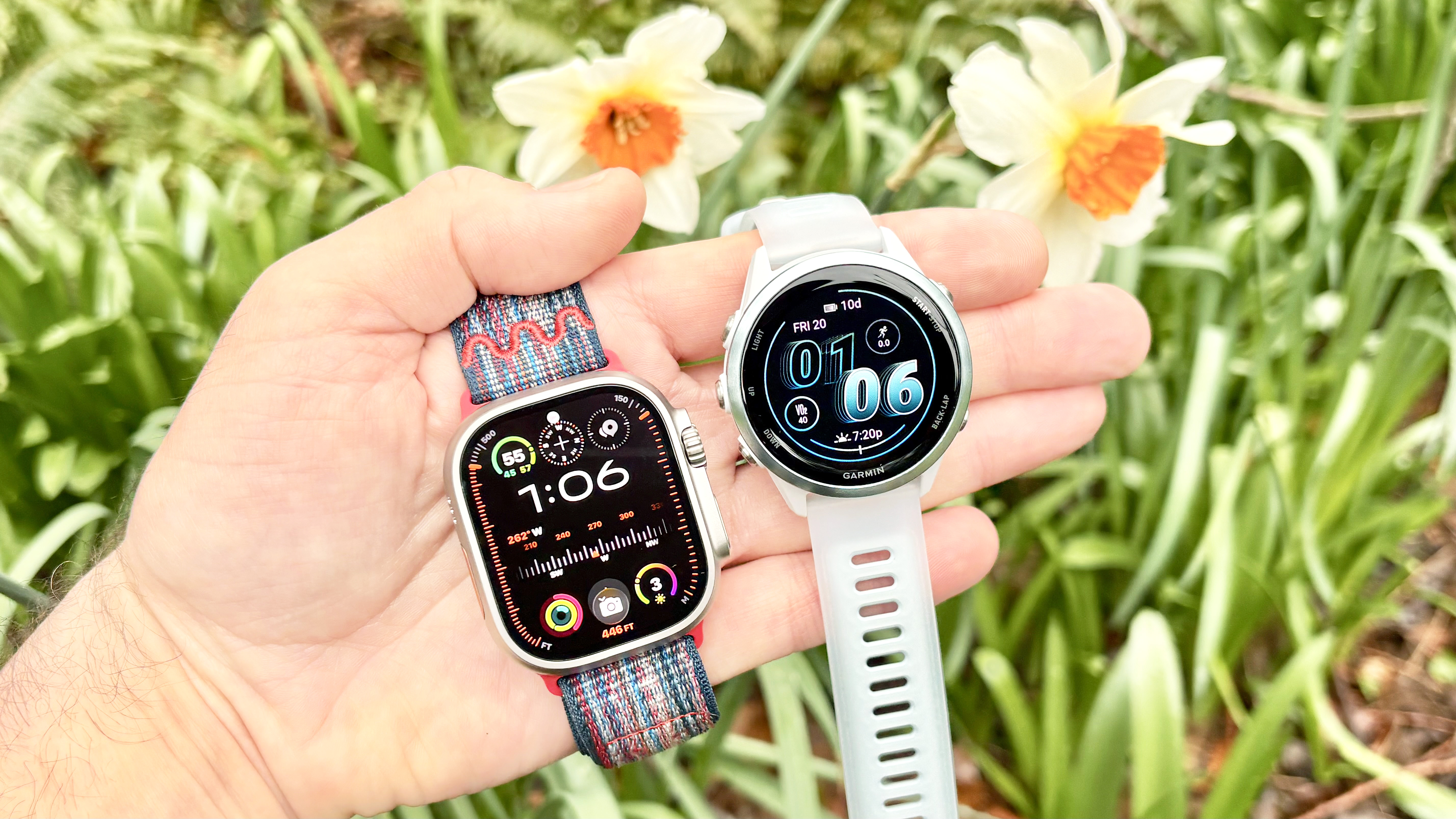 Close-up of the Apple Watch Ultra 3 and Garmin Forerunner 570 in a user's hand with blooming flowers in the background