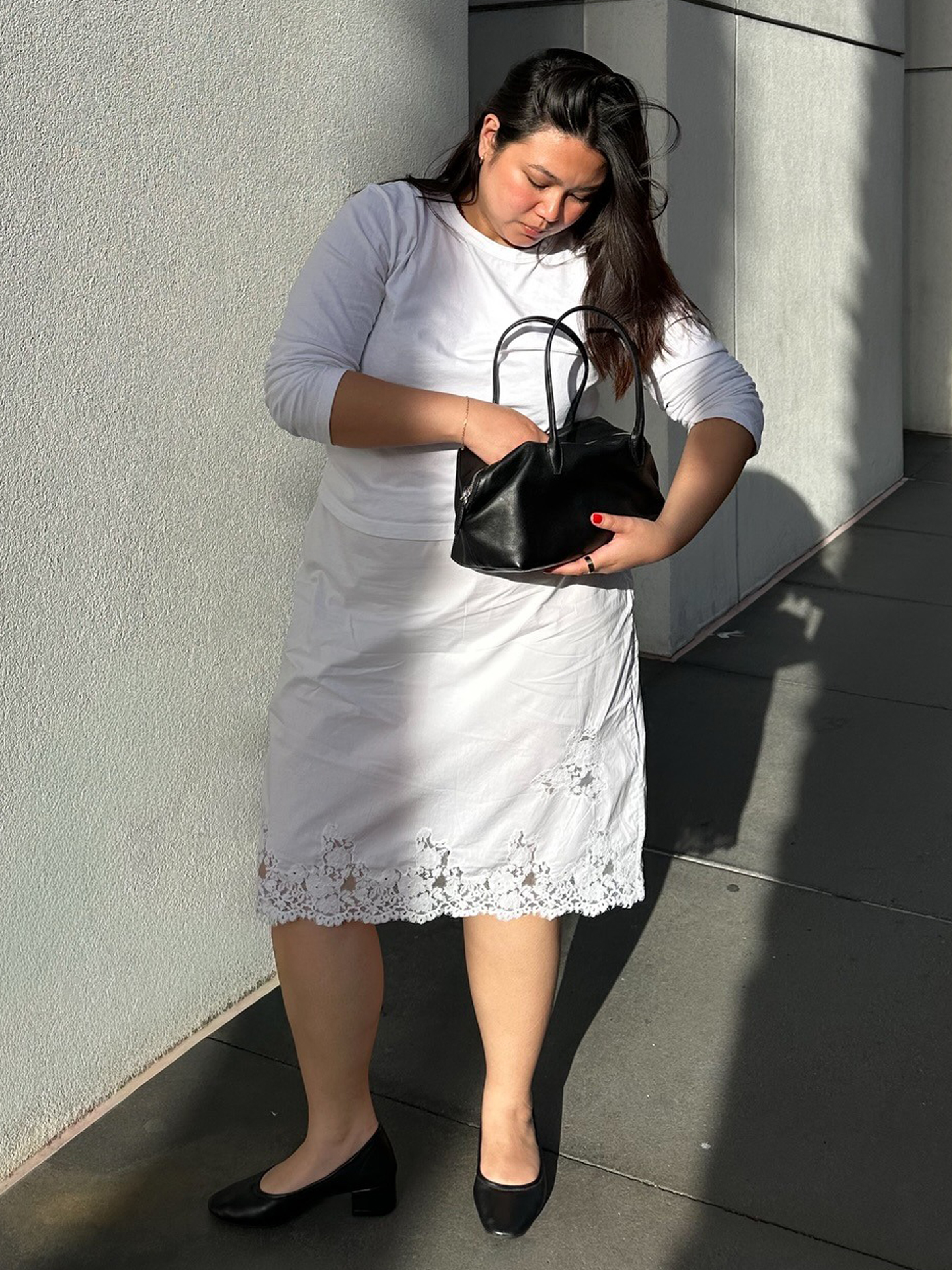 style influencer poses in a long-sleeve white T-shirt, a lace-trim white skirt, and black block heels while looking in a black shoulder bag,