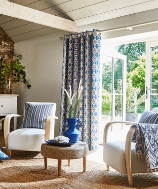 A living room with blue curtains, white chairs with blue pillows, and open french doors leading to sunny leafy garden