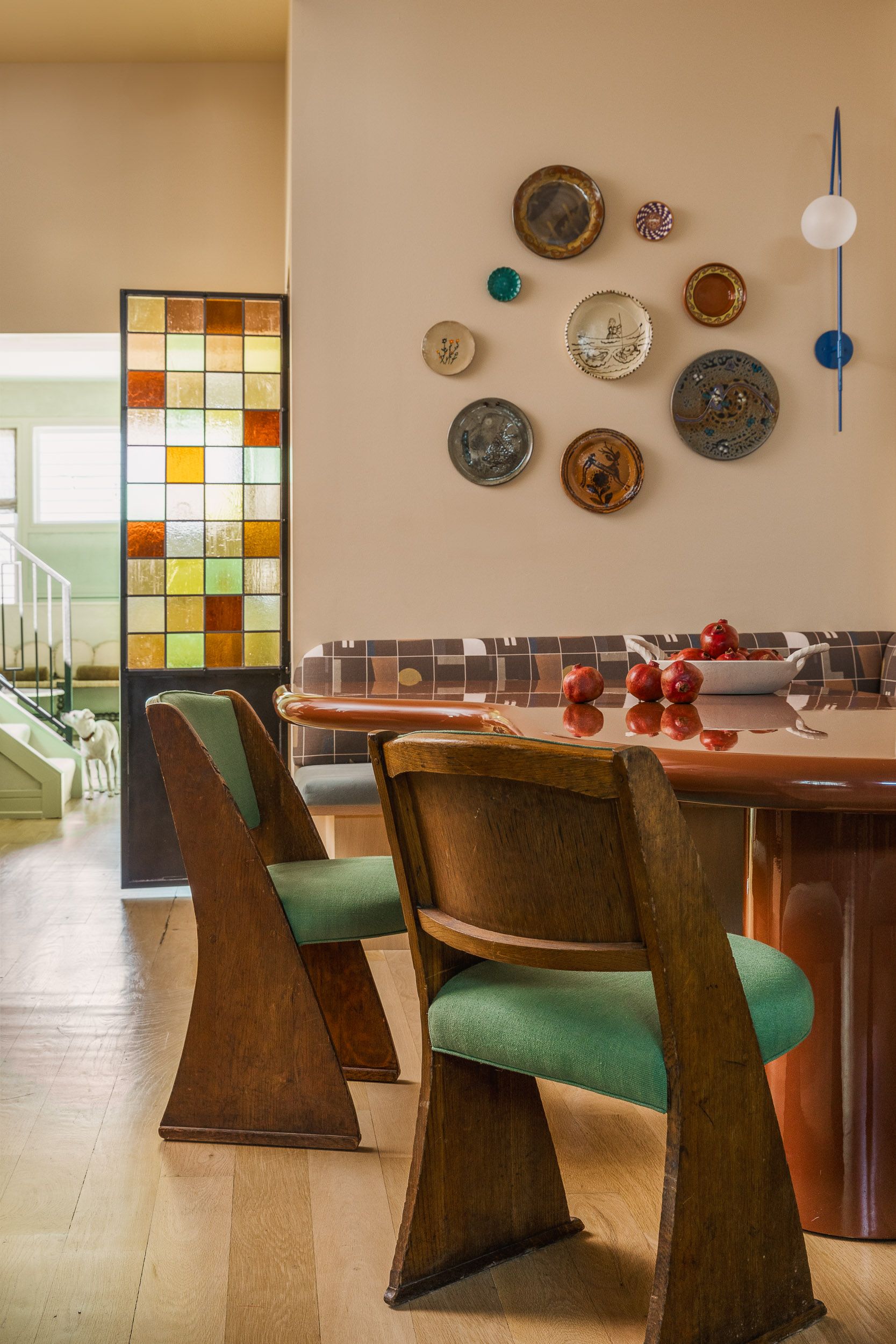 The corner of a dining nook in a light beige kitchen. There is a red lacquer table and two wooden chairs with green seating. There are several plates hung on the wall.