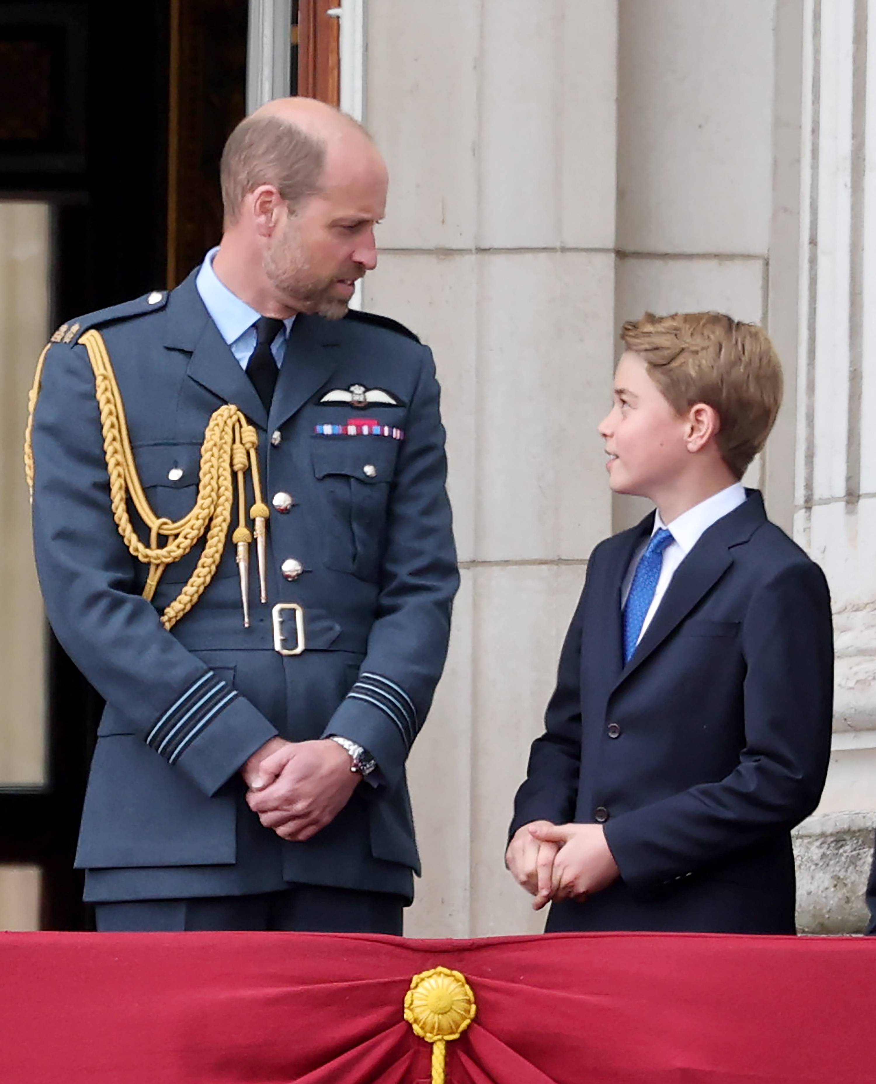 Prince William and Prince George wearing blue suits on the balcony of Buckingham Palace looking at each other