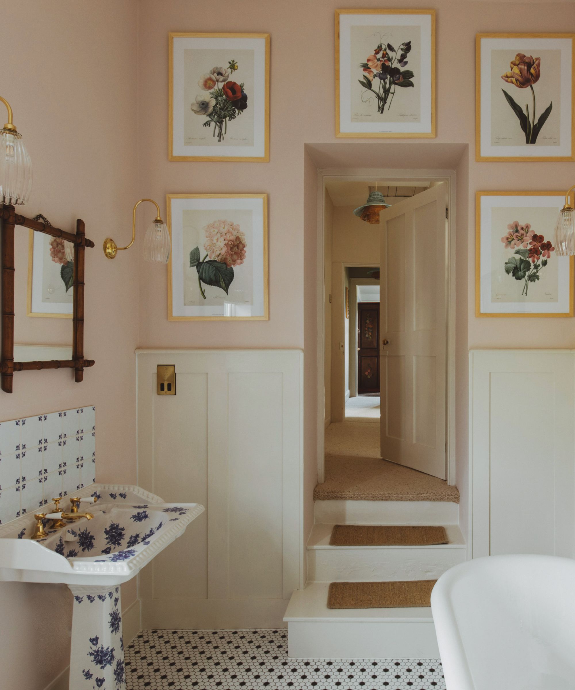 A rustic bathroom with pale pink walls, warm white panelling, floral artworks, black and white flooring, and a blue floral sink.