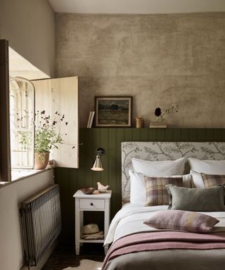 Bedroom with organic, relaxed color palette, double bed with upholstered headboard, green wooden paneling, dark wooden flooring, bed decorated with scatter cushions, throws