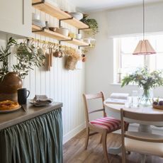 A panelled white kitchen with a dining area featuring a round farmhouse table and open shelving
