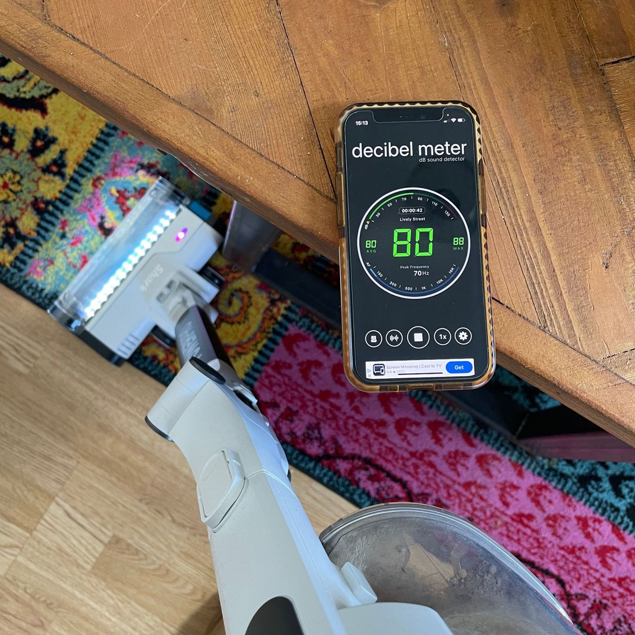 Shark PowerPro Pet Cordless Stick Vacuum on a rug, with a decibel meter app sitting on a table next to it