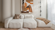 A white living room with a textured white sectional sofa, a framed painting, a structured wall lamp, and a marble coffee table