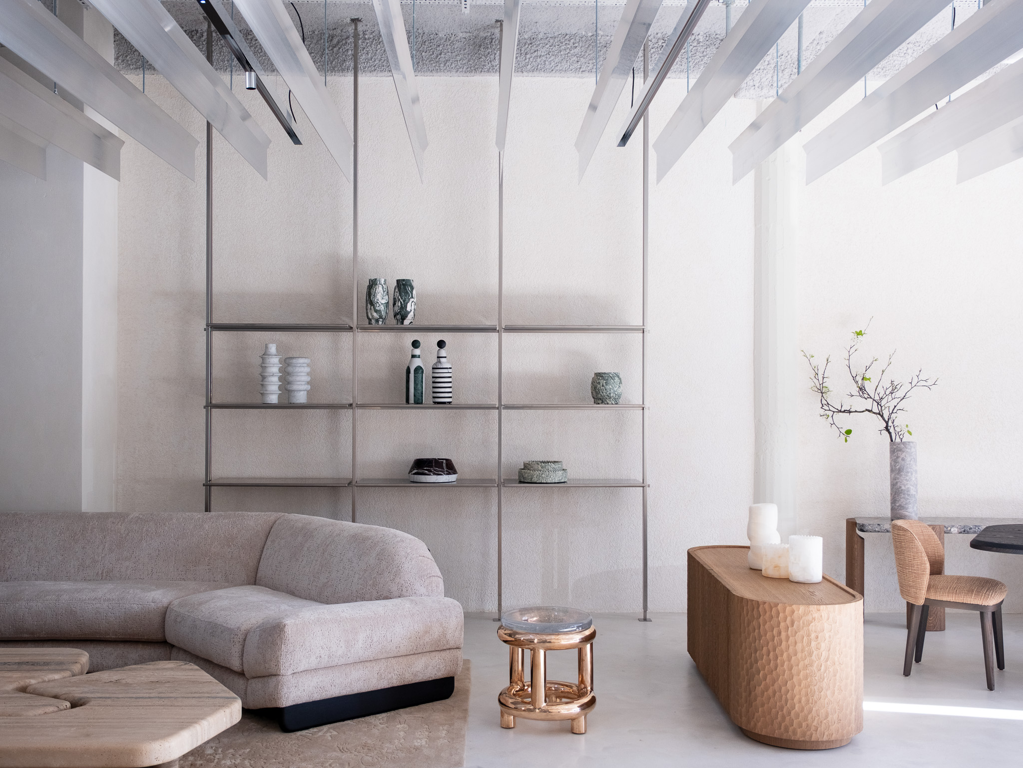 The interior of a brutalist, minial design showroom, decorated with contrasting furniture, artworks, and accessories in wood, earthy fabrics, reflective surfaces, and plants.