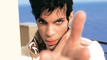 A close-up, color portrait of Prince looking intensely at the camera, with a finger pointing slightly out of focus in the foreground and a blue body of water visible behind him.