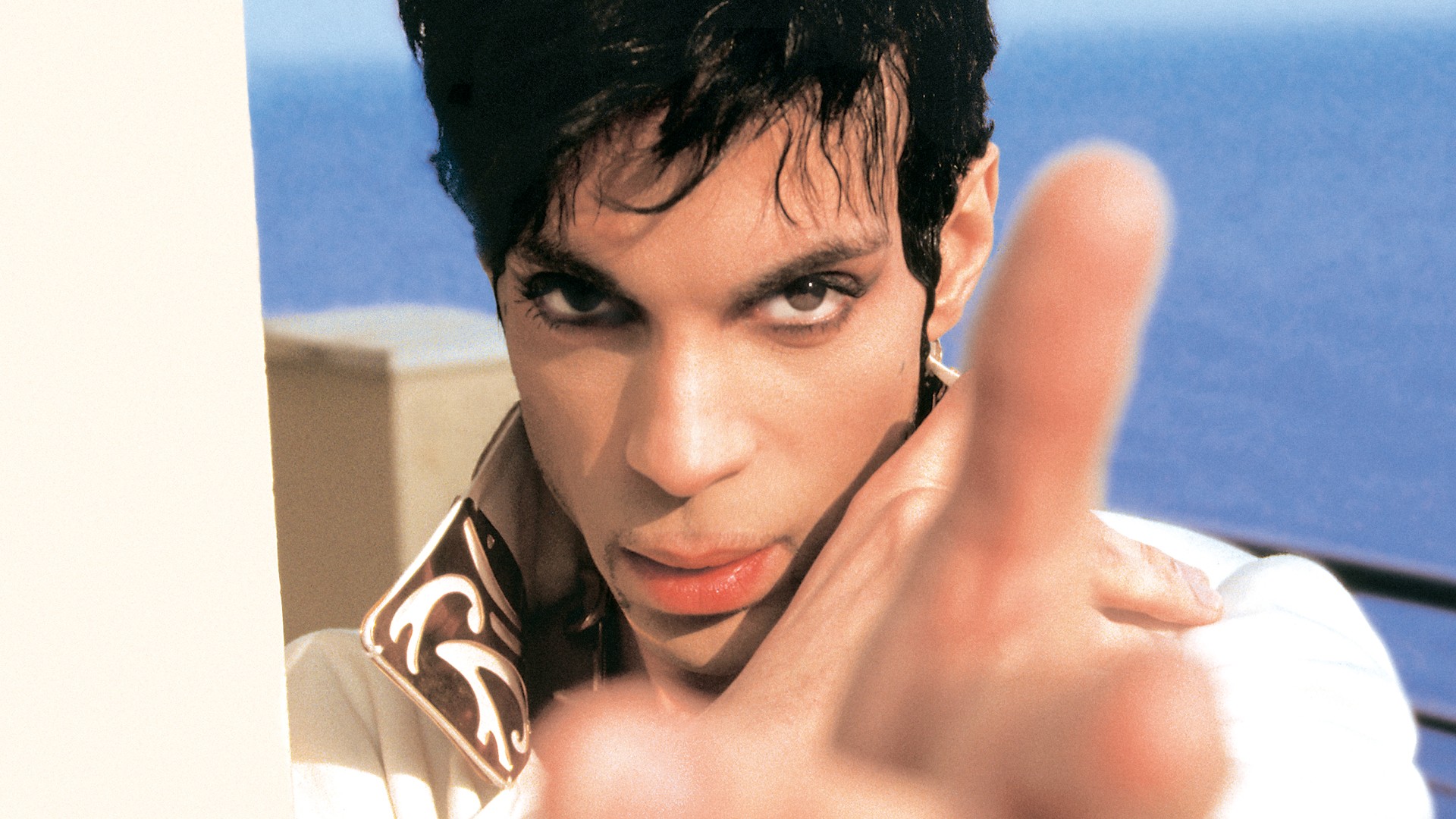 A close-up, color portrait of Prince looking intensely at the camera, with a finger pointing slightly out of focus in the foreground and a blue body of water visible behind him.
