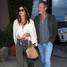 Cindy Crawford walks down the street wearing khaki jeans with beige suede sandals and a white blouse.