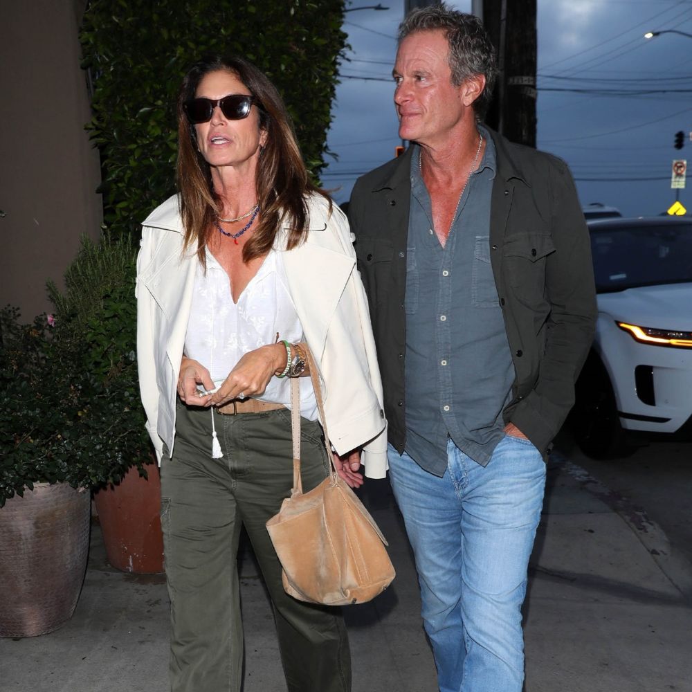 Cindy Crawford walks down the street wearing khaki jeans with beige suede sandals and a white blouse.