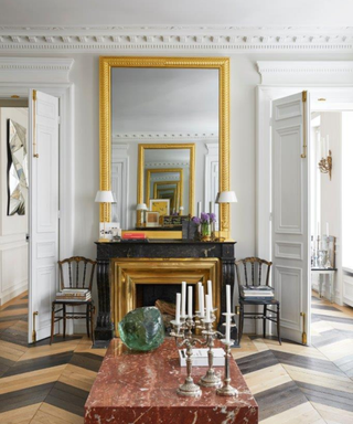 Living room with marble coffee table, gilded mirrors and fireplace framed by wooden dining chairs