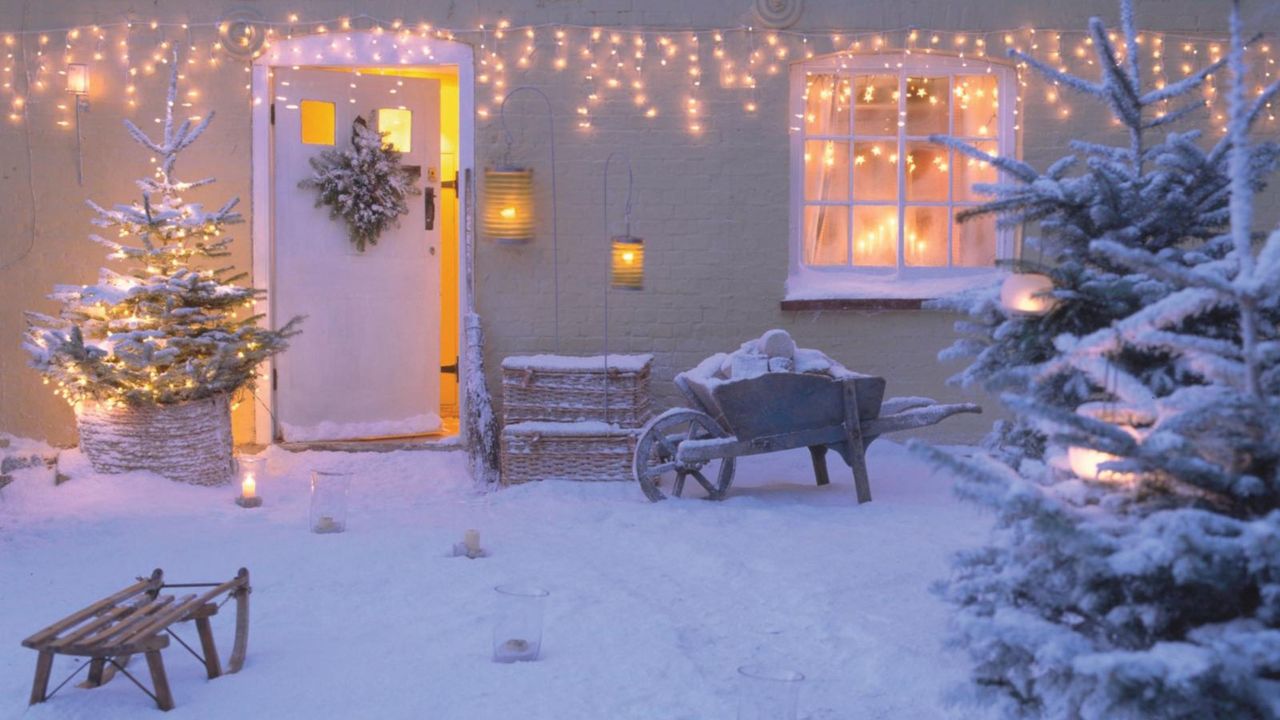 xterior of snow covered house decorated with xmas tree, fairy lights, &amp; lanterns . 