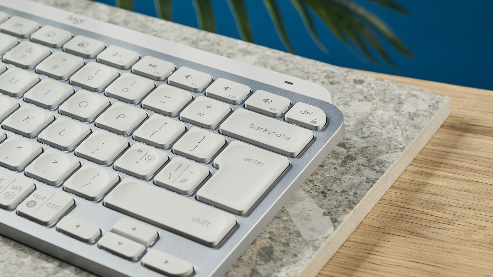 A photograph of the Logitech MX Keys Mini in light gray, positioned on a stone slab with a blue wall in the background.