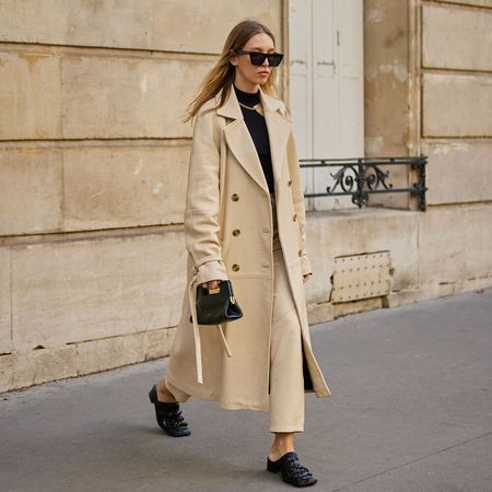 woman wears tan trench coat, tan pants, black sweater, black handbag, and black mule clogs while posing for a street style type of photo