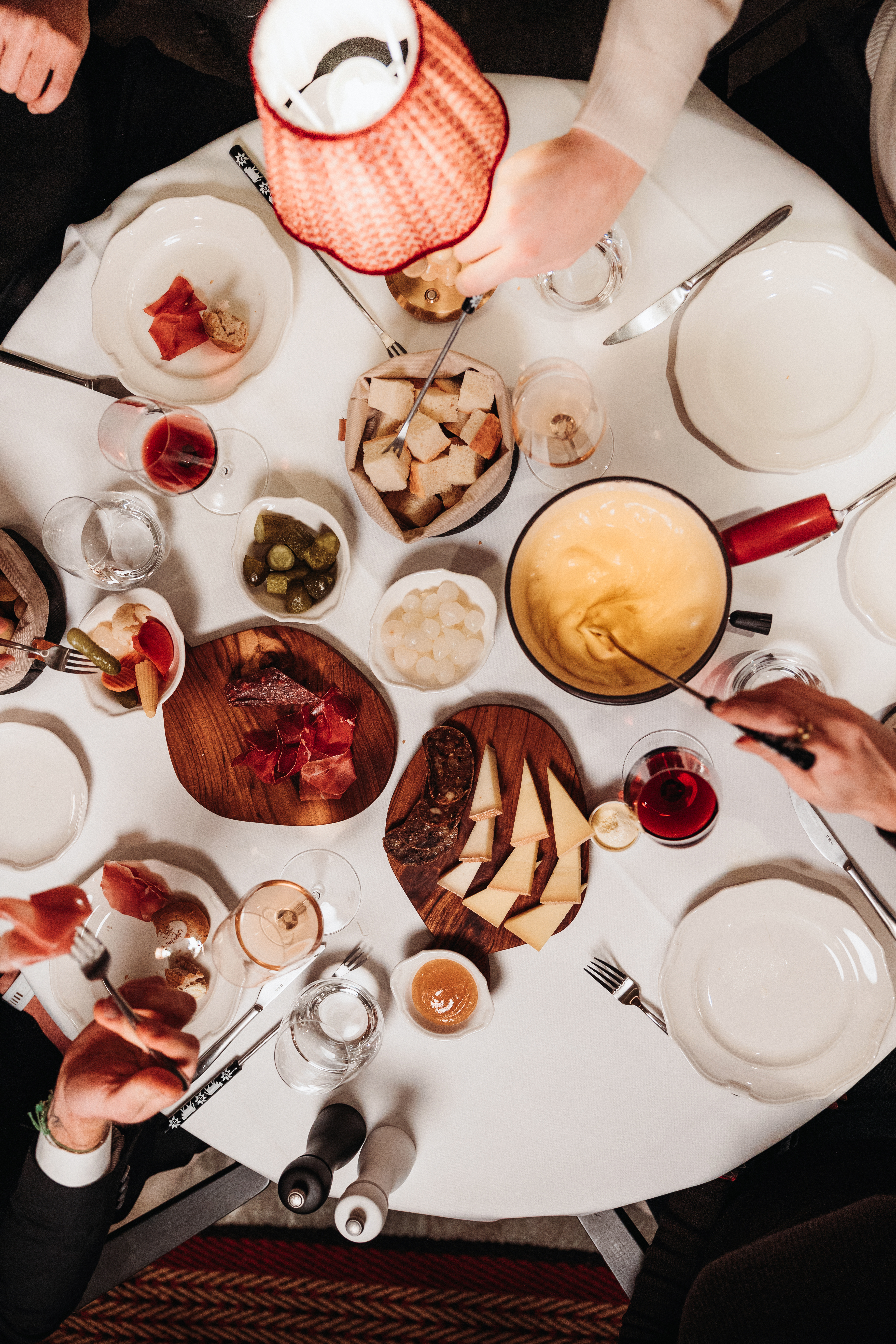 An aerial view of a restaurant table captures a series of hands as they reach to a pot filled with melted cheese, charcuterie, pickles, and wine glasses.