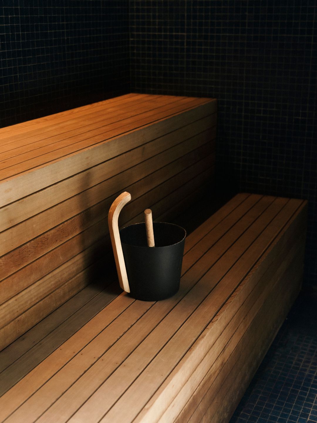 Two angles of a sauna: wooden benches and a bucket with water and ladel
