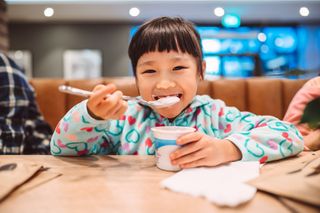 A little girl with dark hair is sat at a table eating a yoghurt with a spoon. She is smiling at the camera.