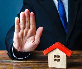 hand of male wearing suit in stop pose with wooden house with red roof on table by his hand