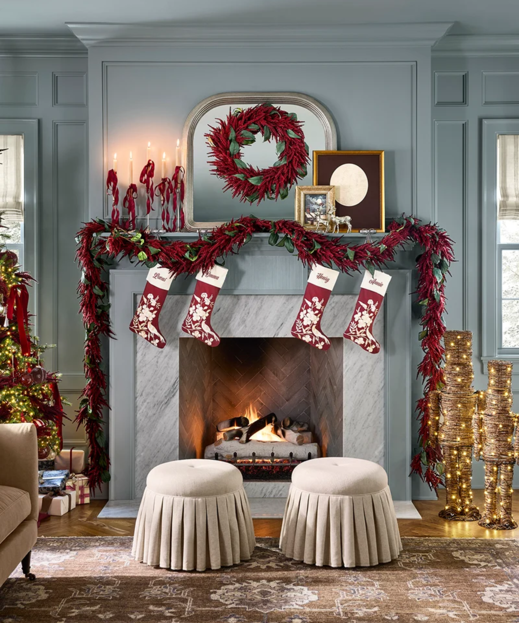 Festive pale blue living room featuring two skirted tan ottomans flanking a roaring marble fireplace decorated with embroidered stockings, Amaranthus garland, and a coordinating wreath.