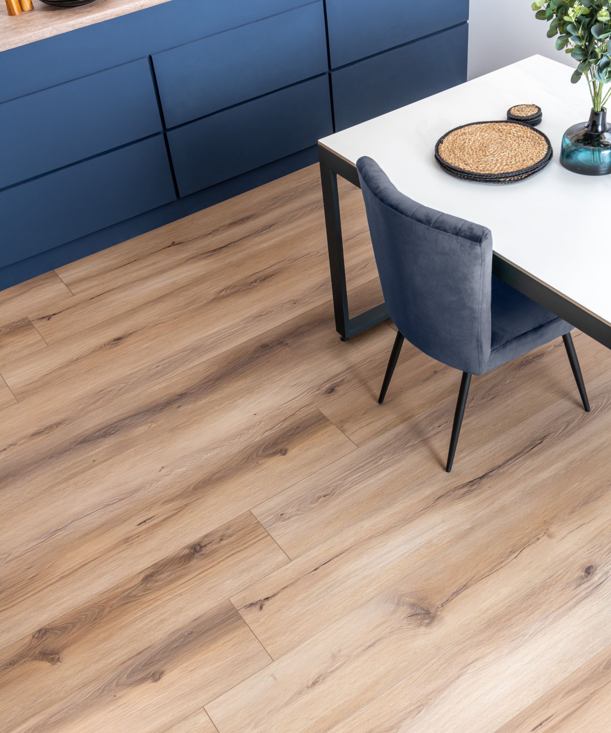 Wide planks in a pale, natural oak tone run lengthways across the space. Subtle knots and grain variations add texture, while the matte finish keeps it soft and understated. The warm beige undertone balances the deep blue cabinetry and cool grey furniture, giving the room an airy, contemporary feel.