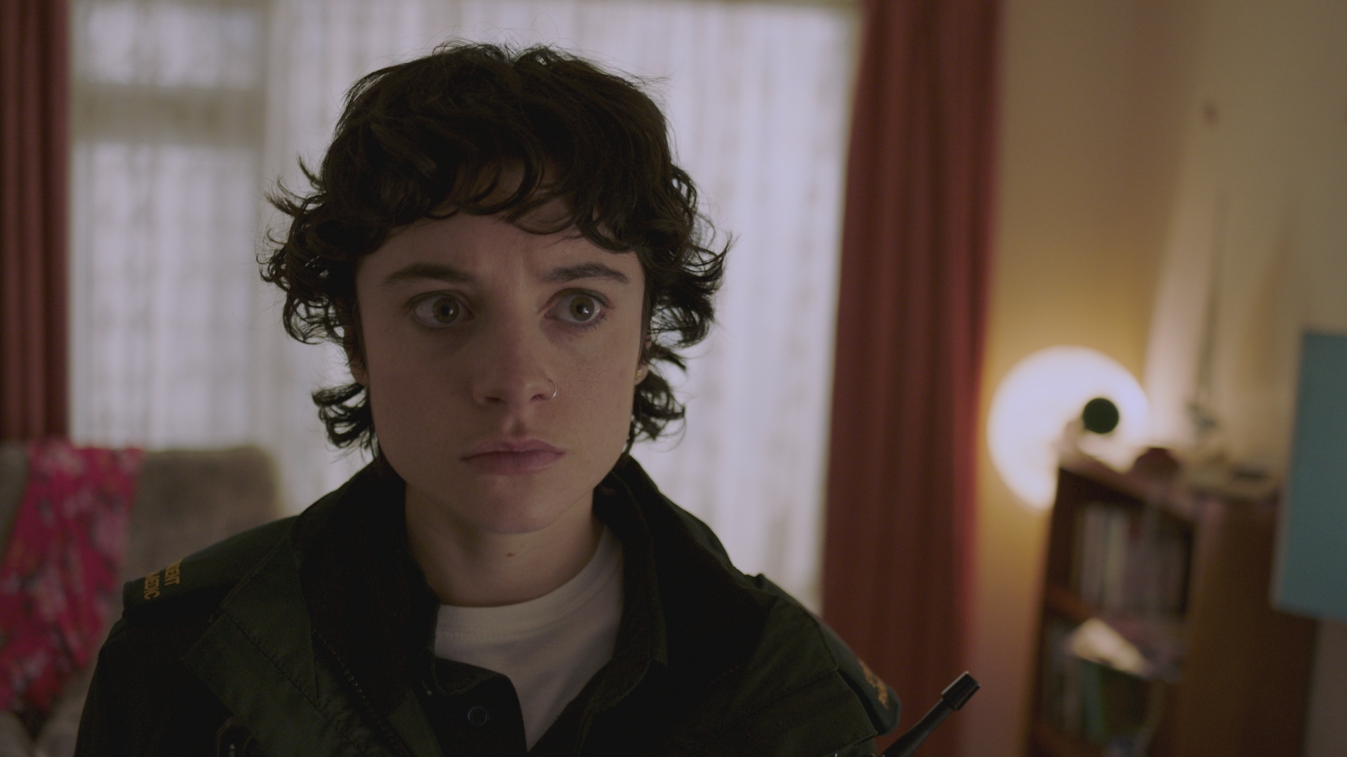 Indie in her paramedic uniform wearing a white t-shirt underneath looking scared inside a dimly lit house.