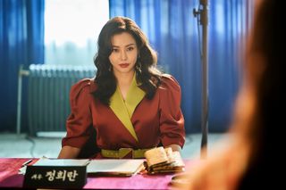 A woman sits at a desk with notebooks and a Korean nameplate, with a radiator behind her, in 'Aema.'