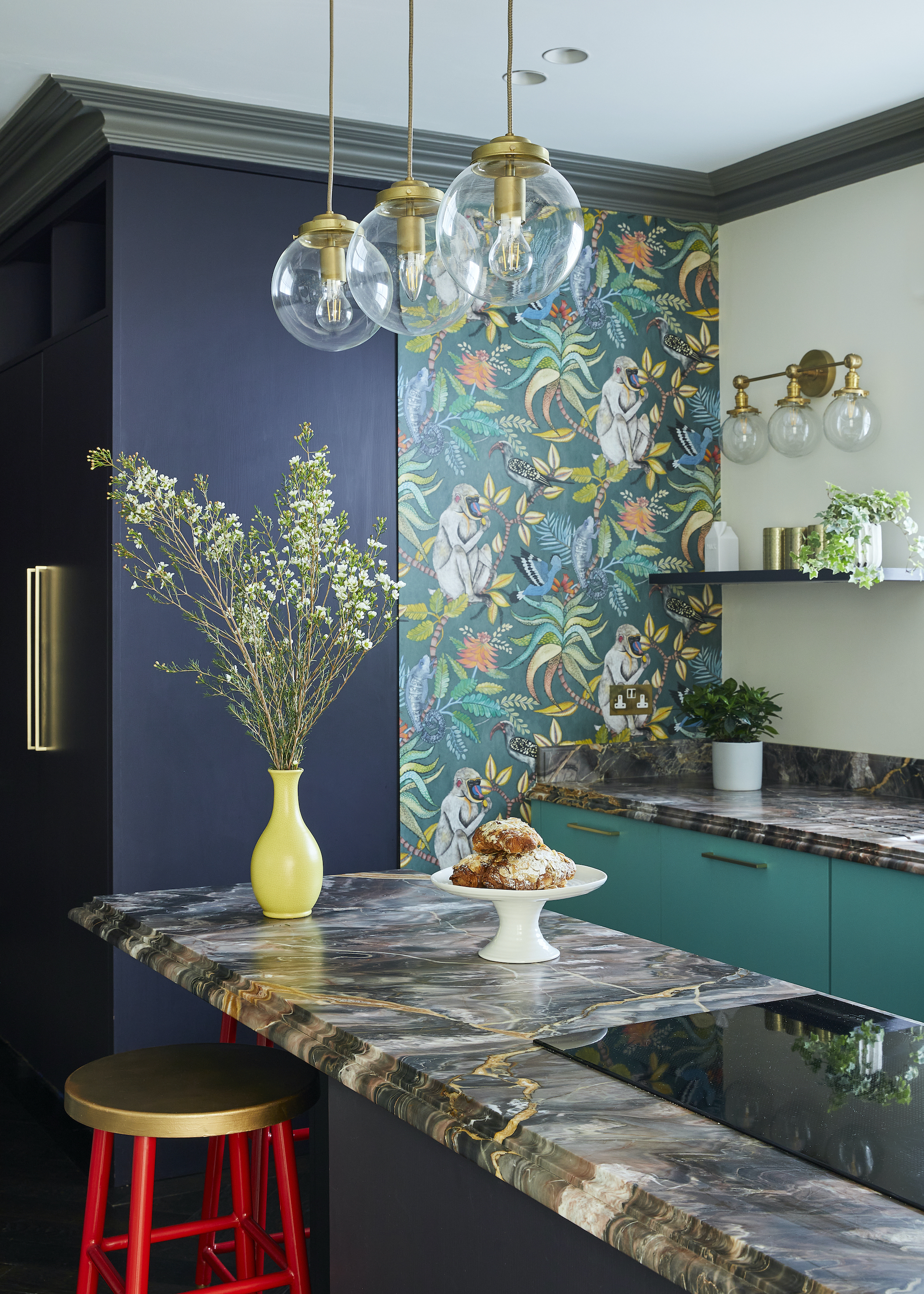 blue kitchen with panel of floral wallpaper with navy blue cabinets, marble counter with red bar stool, yellow vase, teal green cabinets, and white wall with open shelving