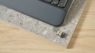 a photo of the 2.4GHz dongle of a black full sized keyboard with batteries and a palm rest on a clean beige surface with a blue backdrop