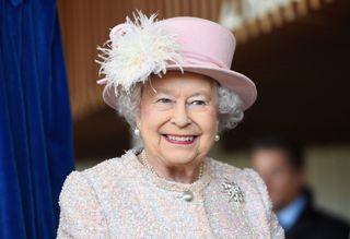 Queen Elizabeth smiling, wearing a pink tweed coat and pink hat with white feathers and pearl earrings