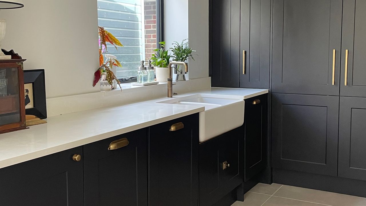 Corner of a dark grey kitchen with tall units on one side and butler sink on a white worktop under the window