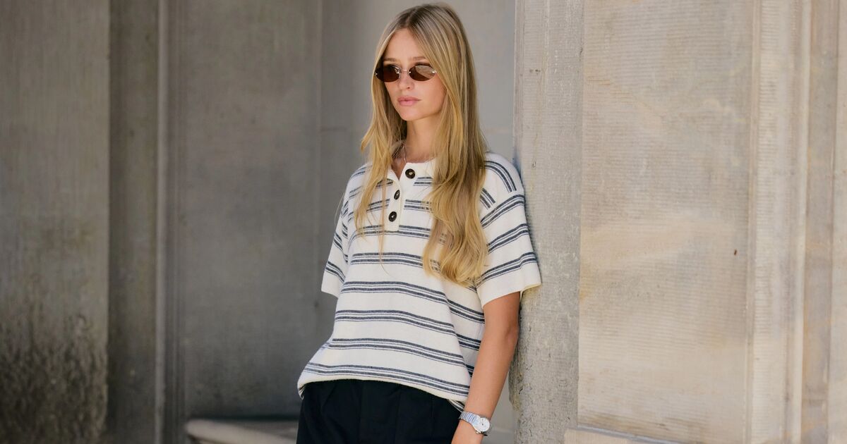 Woman wears striped top, black mini skirt, pointed toe heels, and a white handbag while leaning on a wall. 