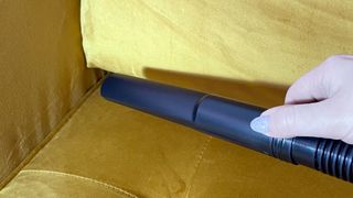 A close-up of the nozzle attachment and extension hose, held by a woman's hand over a mustard colored sofa.