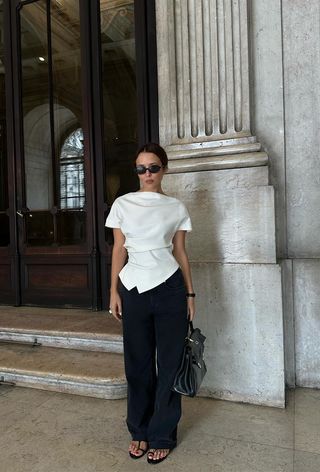 a photo showing what shoes to wear with wide-leg pants with a woman standing in front of a door wearing a white draped blouse with black wide leg linen trousers and black strappy flat sandals and a black handbag