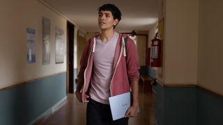 A young person in a red hoodie holds a laptop and notebook while walking down a hallway with light-colored walls and posters.