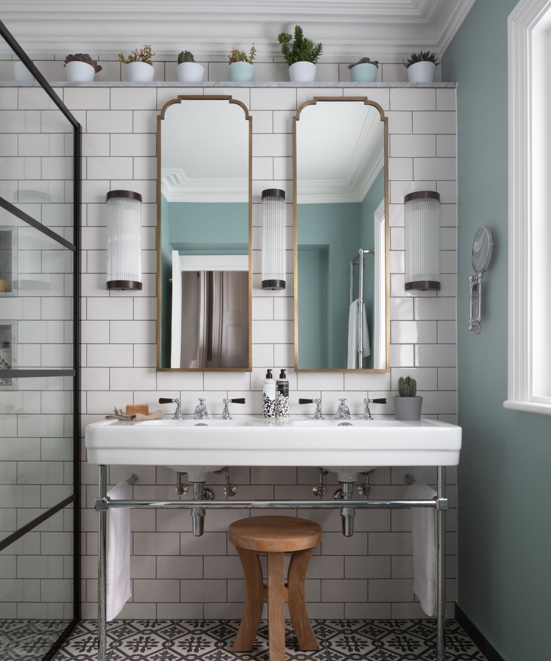 A white bathroom with gold mirrors, patterned floor tiles, and house plants on a high shelf