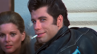 John Travolta as Danny Zuko looking at camera with leather jacket on and cigarette in his mouth as he smiles in Grease