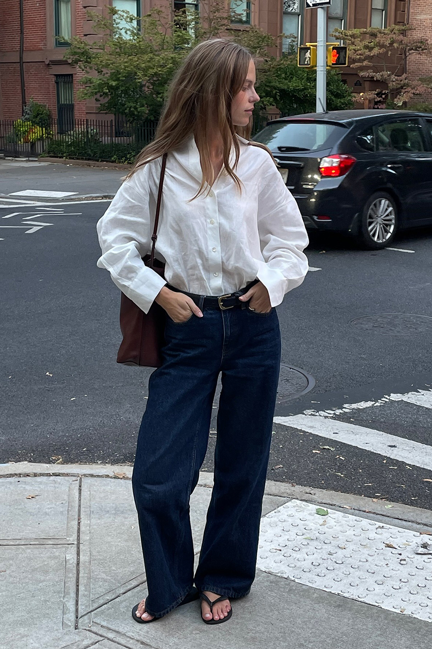 a woman wearing a white button-down, blue jeans, burgundy bag, and black flip flops